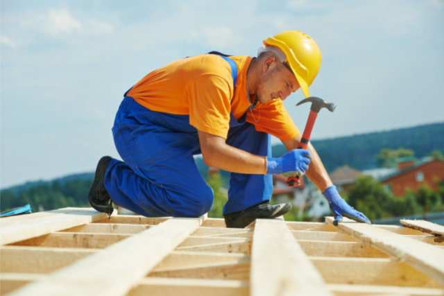 Re-roofing by worker with a yellow hard hat on and blue gloves with a hammer.